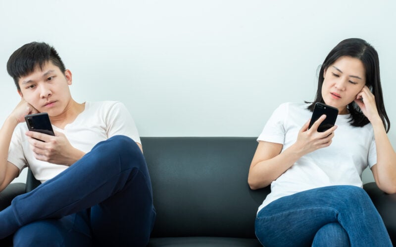 a young couple sits on a couch but are looking at their phones