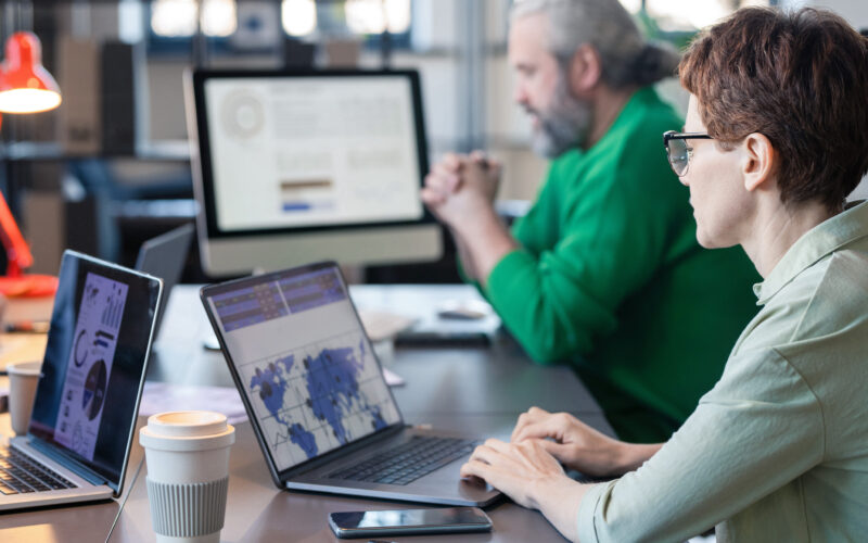 Rear view of businesswoman in eyeglasses sitting at the table and typing on laptop, she analyzing charts online with her colleagues in the background